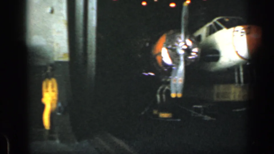 Dim industrial or engine-room machinery appears in a grainy 8mm frame, with metal components and a bright yellow object at left.