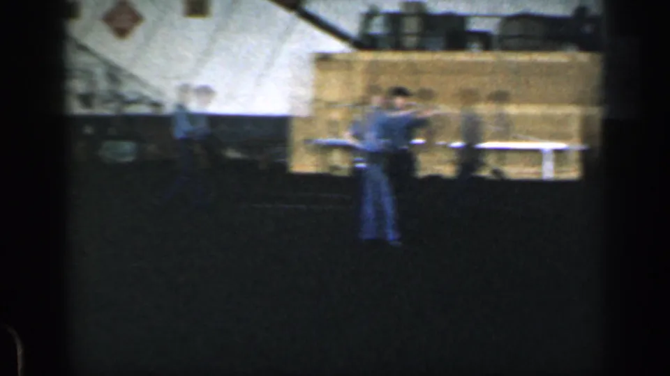 A uniformed man stands in a shadowed Cuban warehouse area with stacked cargo or containers behind him.