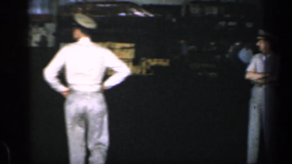 Two men in light uniforms are seen in a dim 1962 Cuba warehouse, one facing away from the camera.