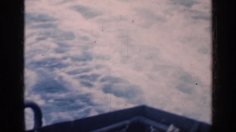 Ocean wake spreads behind the moving ship, with dark railing and 8mm film texture visible in the foreground.