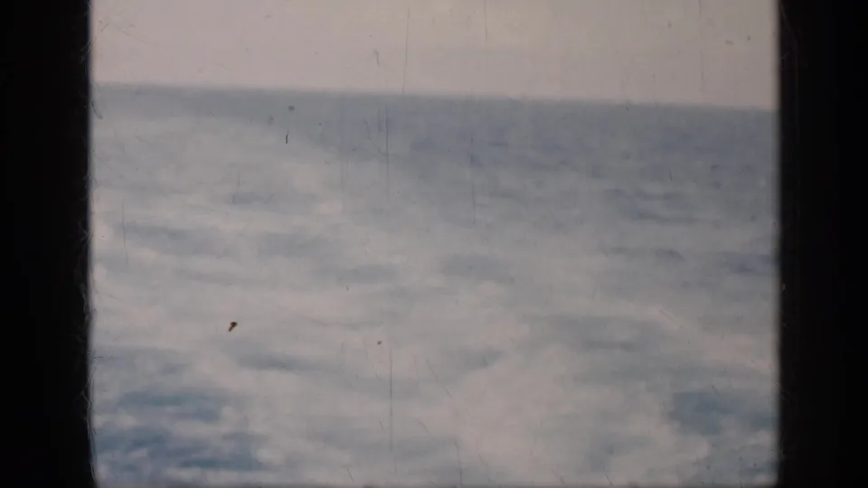 1962 Cuba 8mm home-movie frame showing rough blue-gray sea under a pale cloudy sky, viewed through dark window bars.