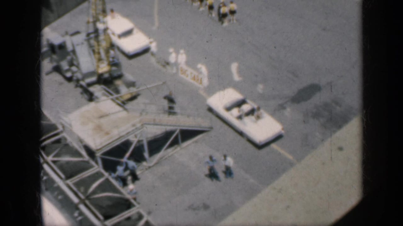 Elevated view of paved industrial area with white cars, crane parts, and small groups of people.