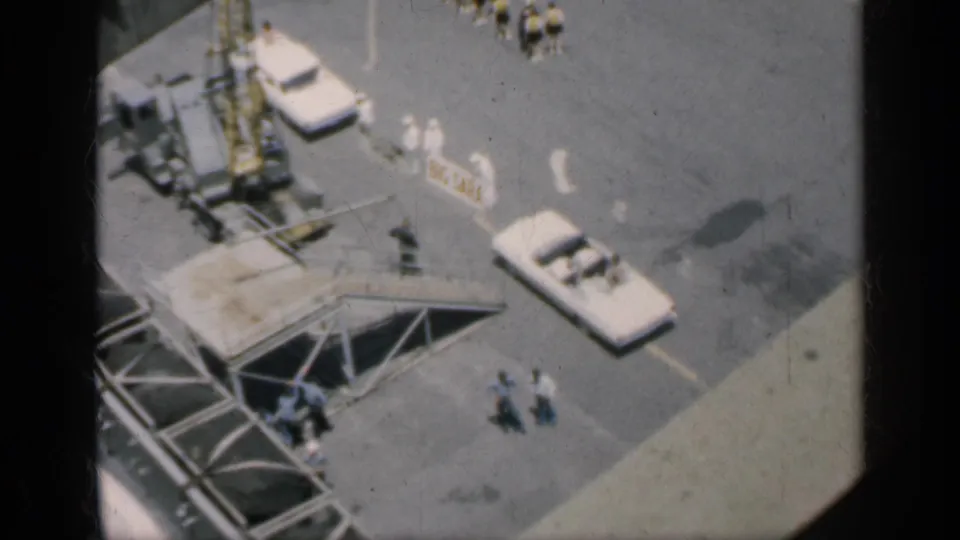 1962 Cuba 8mm frame looking down over a paved work area with white cars, crane equipment, shadows, and scattered people.