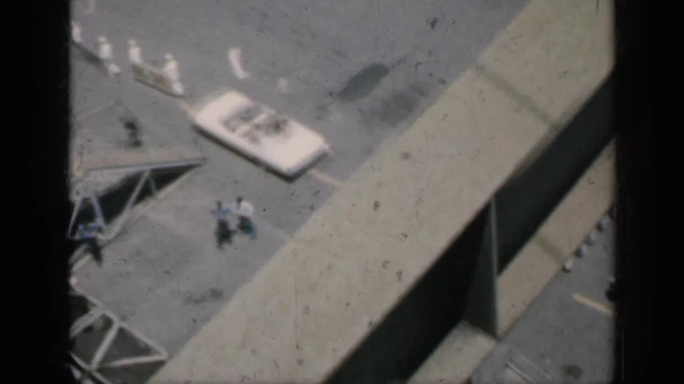 1962 Cuba frame with a large foreground beam, white cars, and small figures moving across a paved industrial area.
