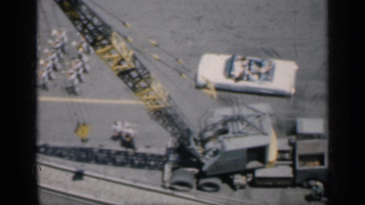 Crane, vintage car, sailors, and deck equipment on an aircraft carrier deck.