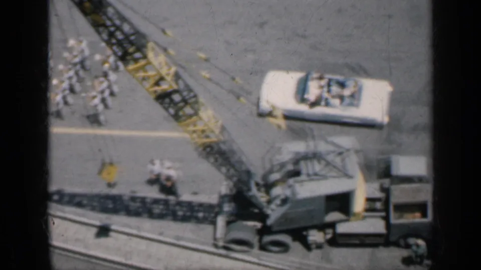 Cuba-linked 1962 8mm frame showing a carrier deck with a crane, a vintage car, sailors in white, and equipment viewed from above.