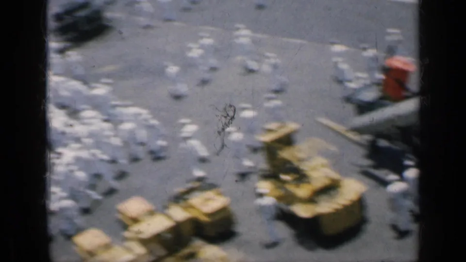 Cuba-linked 1962 8mm frame showing sailors in white near cargo pallets and deck equipment on a carrier deck.