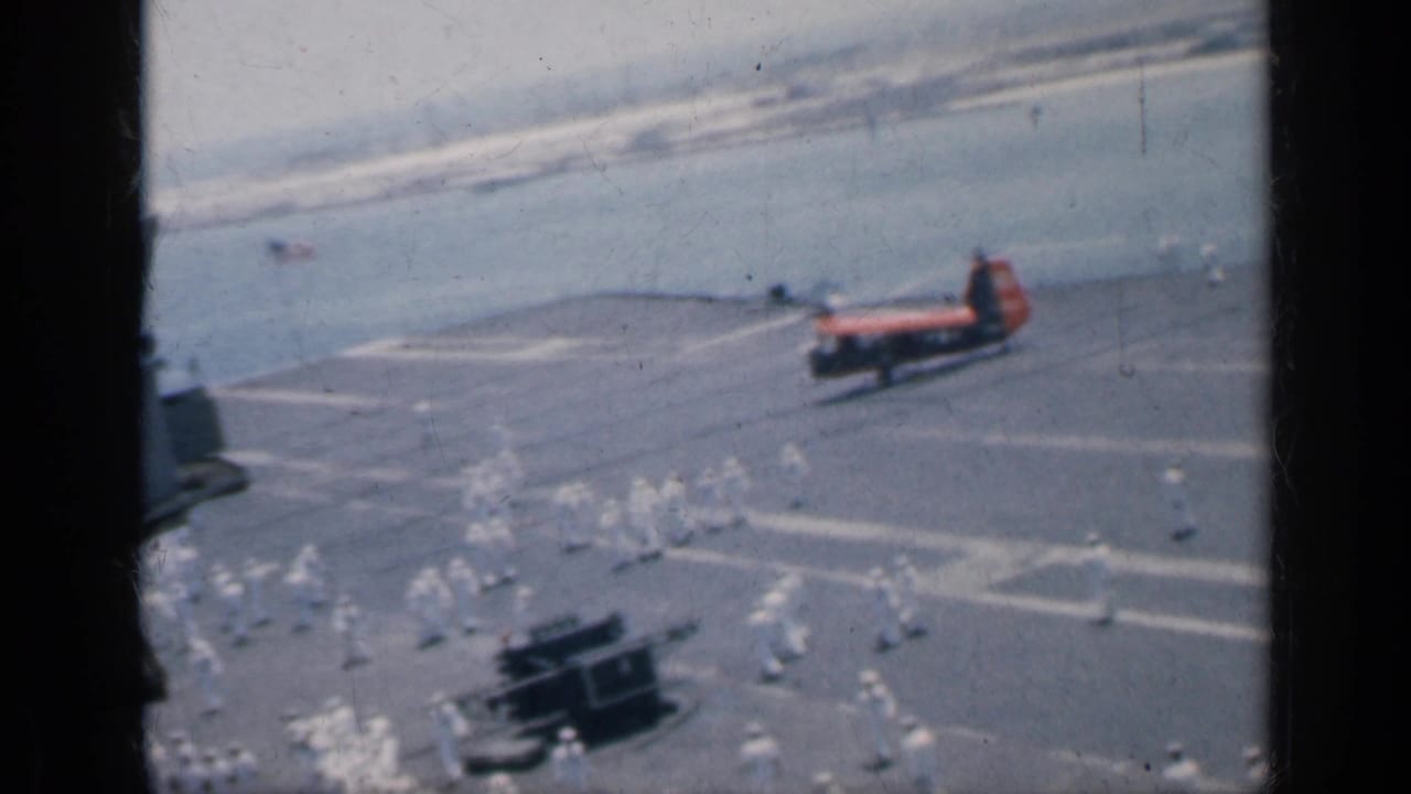 Carrier deck seen through dark window framing, with white-uniformed personnel and a red-orange helicopter near the water.
