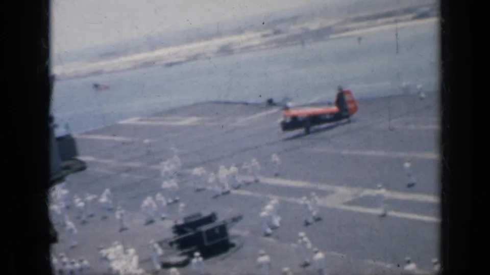 1962 Cuba naval home-movie frame showing an aircraft carrier deck, small groups of personnel in white, and a red-orange helicopter near open water.