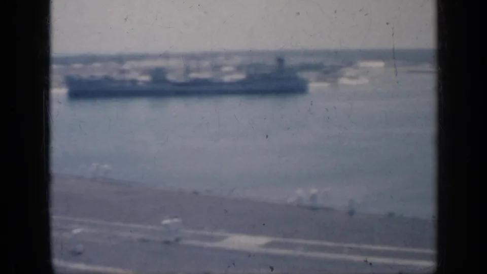 1962 Cuba naval home-movie frame looking past the carrier deck toward a large distant vessel on blue water.