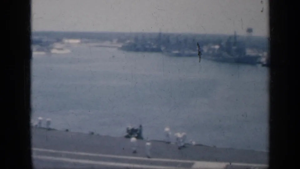 1962 Cuba frame showing a wide view from the aircraft carrier deck toward water, distant ships, and faint shoreline or harbor structures.