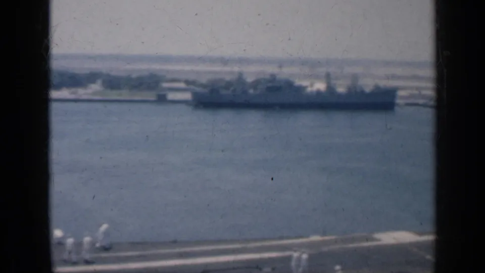 A docked ship appears across the water while crew in white uniforms stand on the filming vessel's deck.