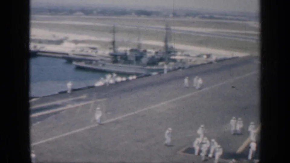 Aircraft carrier deck in Cuba with small groups of naval crew in white uniforms and warships visible across the harbor.