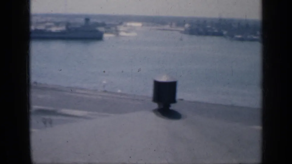 Aircraft carrier deck edge in Cuba overlooking harbor water, distant vessels, and low shoreline through soft 8mm focus.