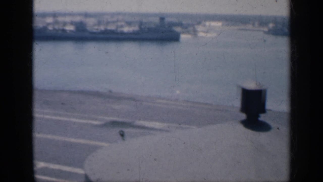 High-angle carrier deck view with harbor water, distant ship, and a dark vertical window or frame edge.