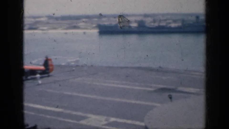 Small military helicopter appears on the 1962 Cuba carrier deck, with harbor water, distant ship, deck markings, and soft port background visible.