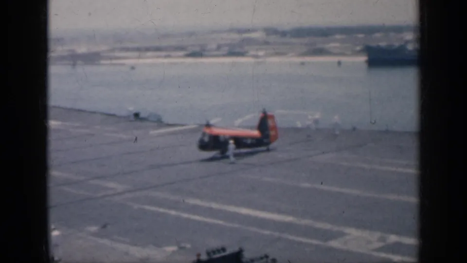 A helicopter sits on the carrier deck in Cuba, 1962, with at least one crew member nearby, harbor water behind it, and a large ship in the distance.
