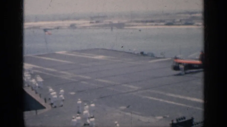 White-uniformed crew march in formation along the 1962 Cuba carrier deck as a small helicopter sits near the frame edge and harbor water fills the background.