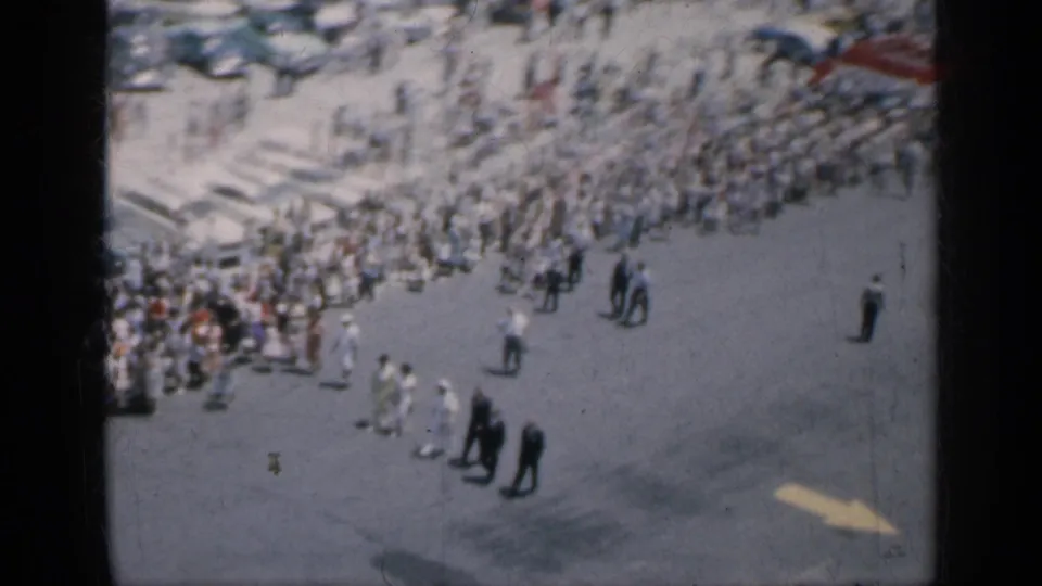 1962 Cuba 8mm frame showing a large crowd gathered near open pavement, with pedestrians crossing the foreground and beach activity beyond.