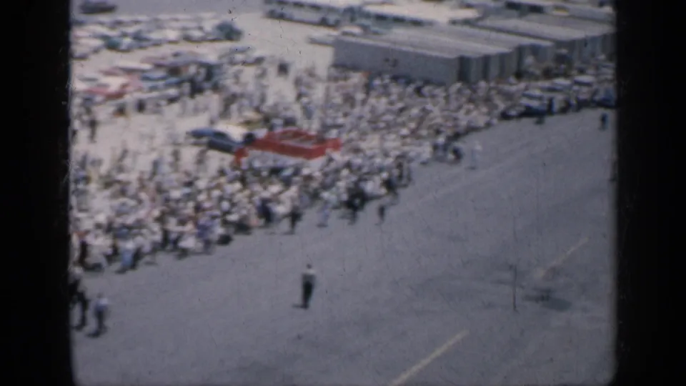 1962 Cuba 8mm frame with dense crowds near parked vehicles and low structures, viewed across a wide paved area under bright daylight.