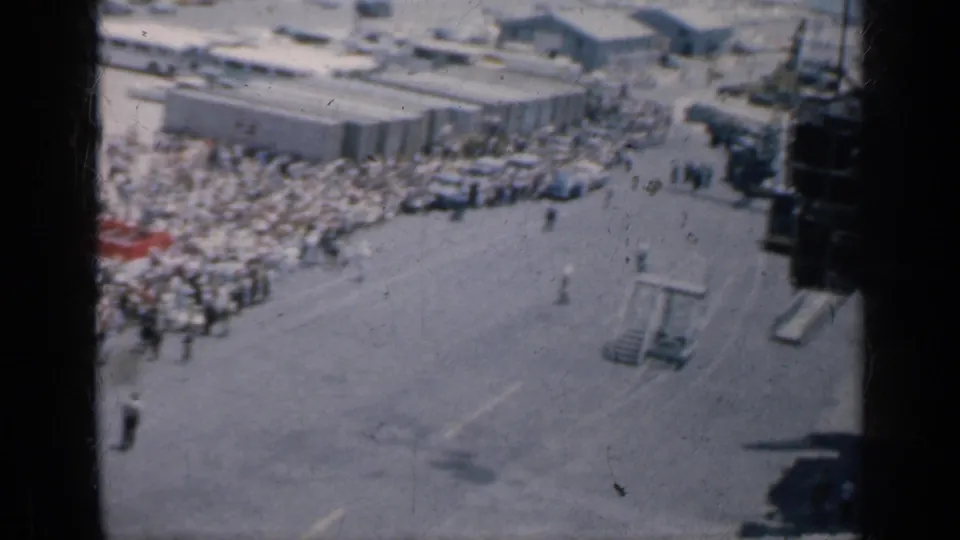1962 Cuba 8mm frame showing a crowd beside a low building, with open pavement, pedestrians, and small structures in the foreground.