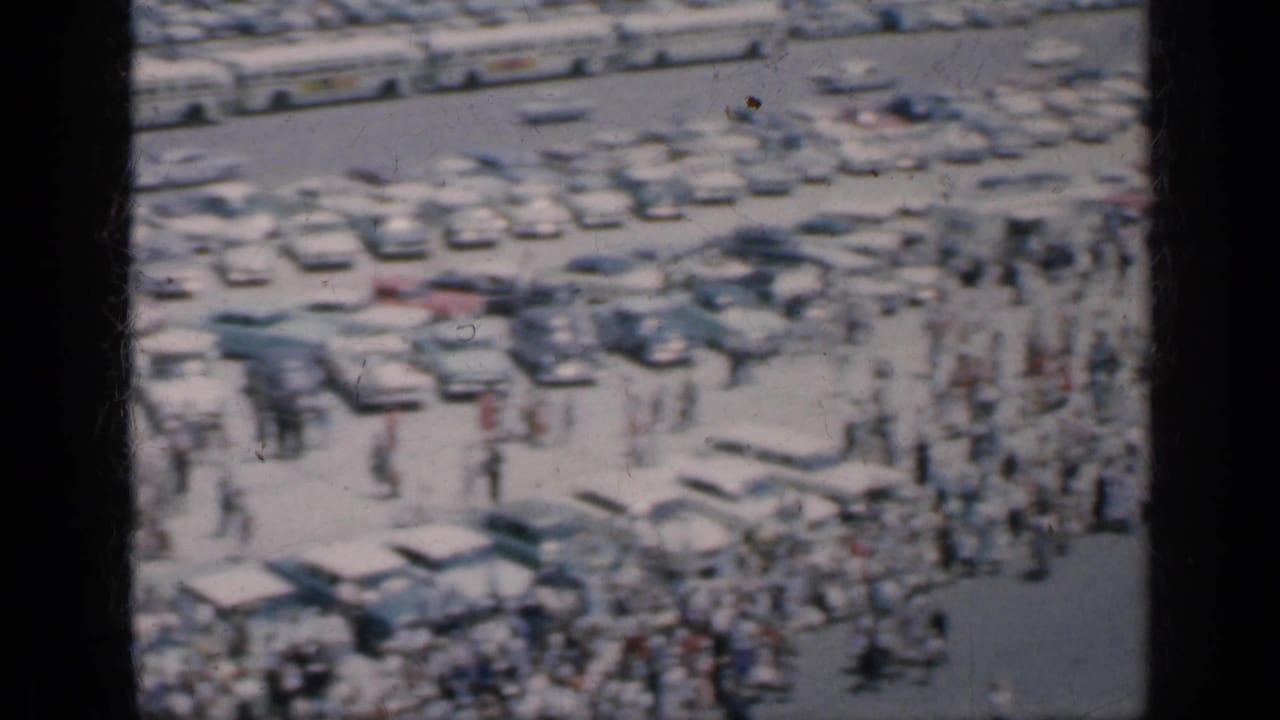 Rows of parked cars and scattered people in a bright outdoor gathering space.