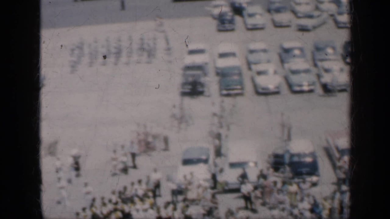 1962 Cuba home-movie frame with people crossing an open paved area near parked cars and a larger crowd below.