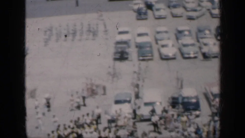 1962 Cuba home-movie frame with people crossing an open paved area near parked cars and a larger crowd below.