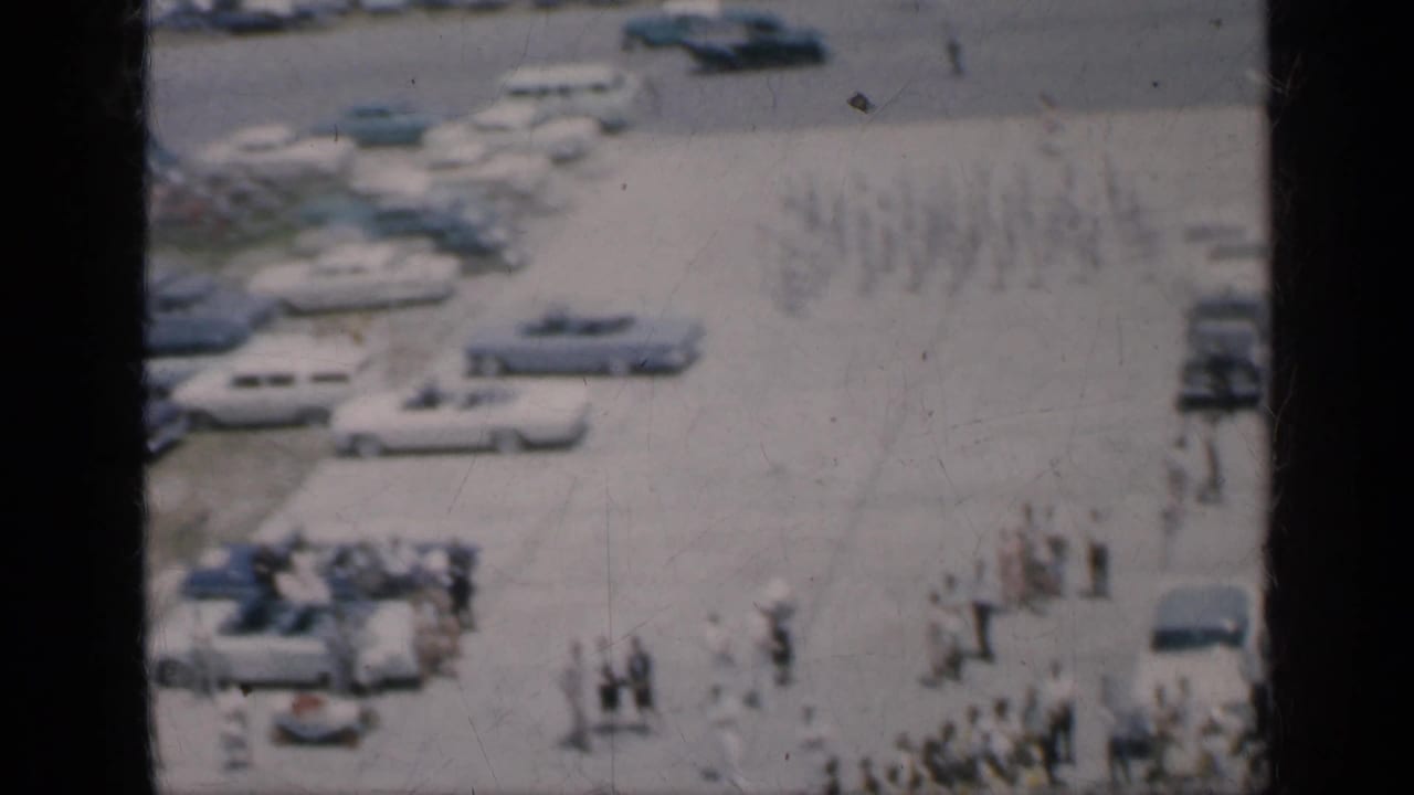 Elevated view of rows of parked cars and small groups of people beside a roadway near water.