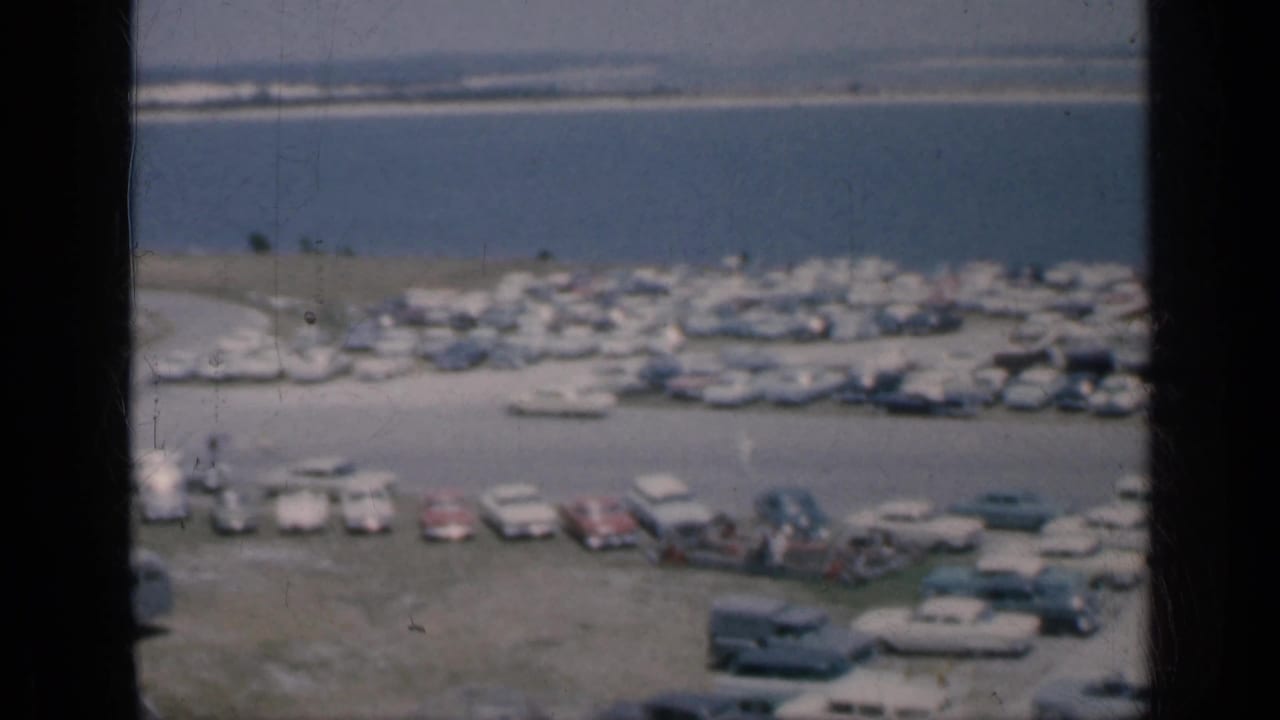 Aerial view of parked cars lined along a waterfront with water and a distant bank behind them.