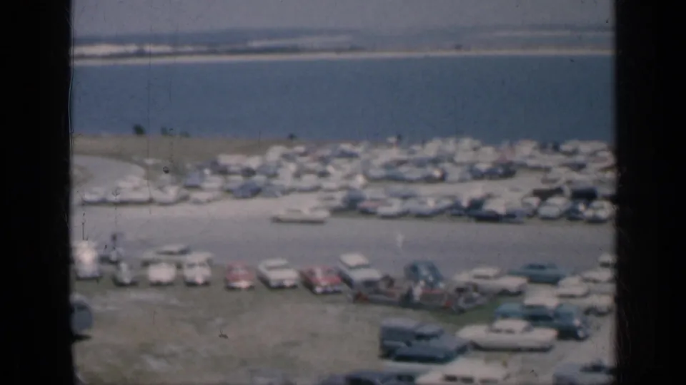 1962 Cuba home-movie frame looking down on rows of cars by a waterfront, with small people visible and a distant shoreline beyond.