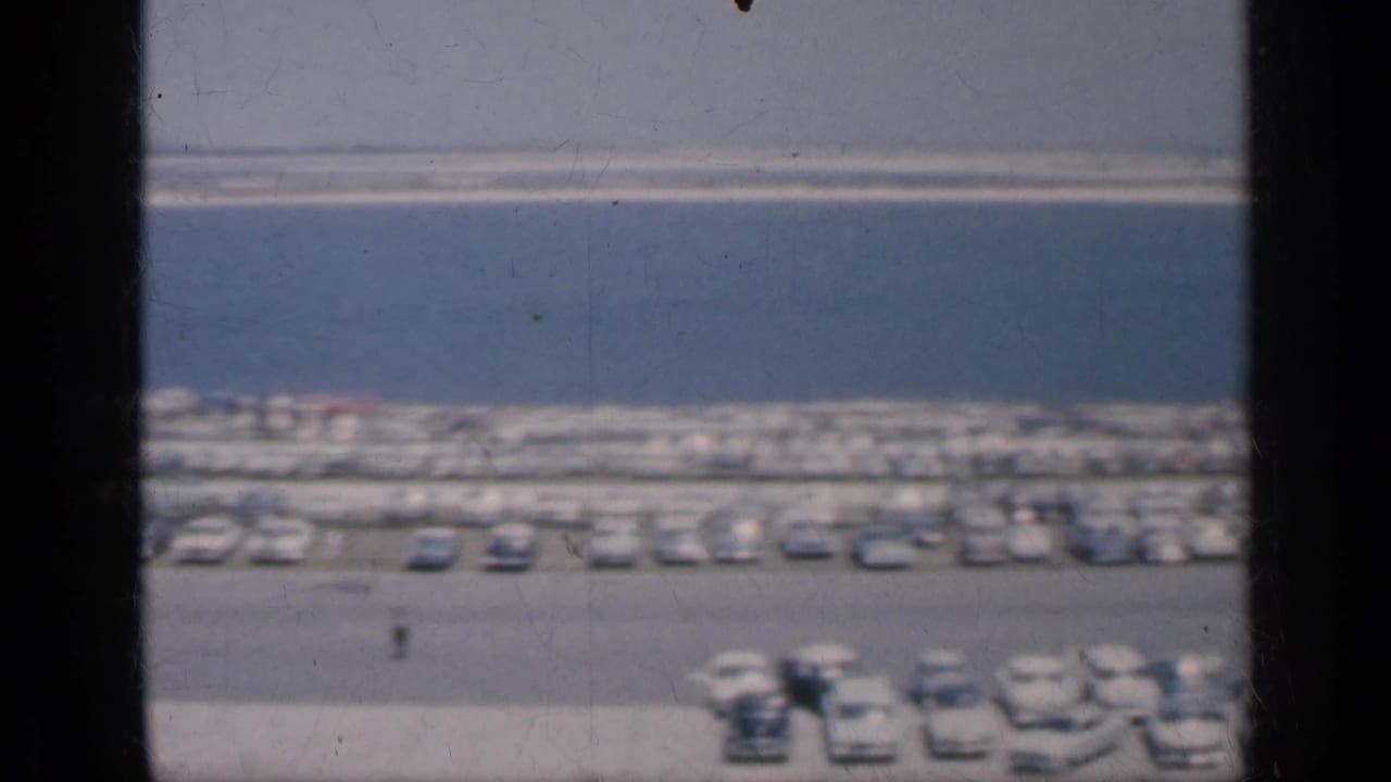 1962 Cuba home-movie frame showing a long line of parked cars near blue water, with a pale bank in the distance and small figures below.
