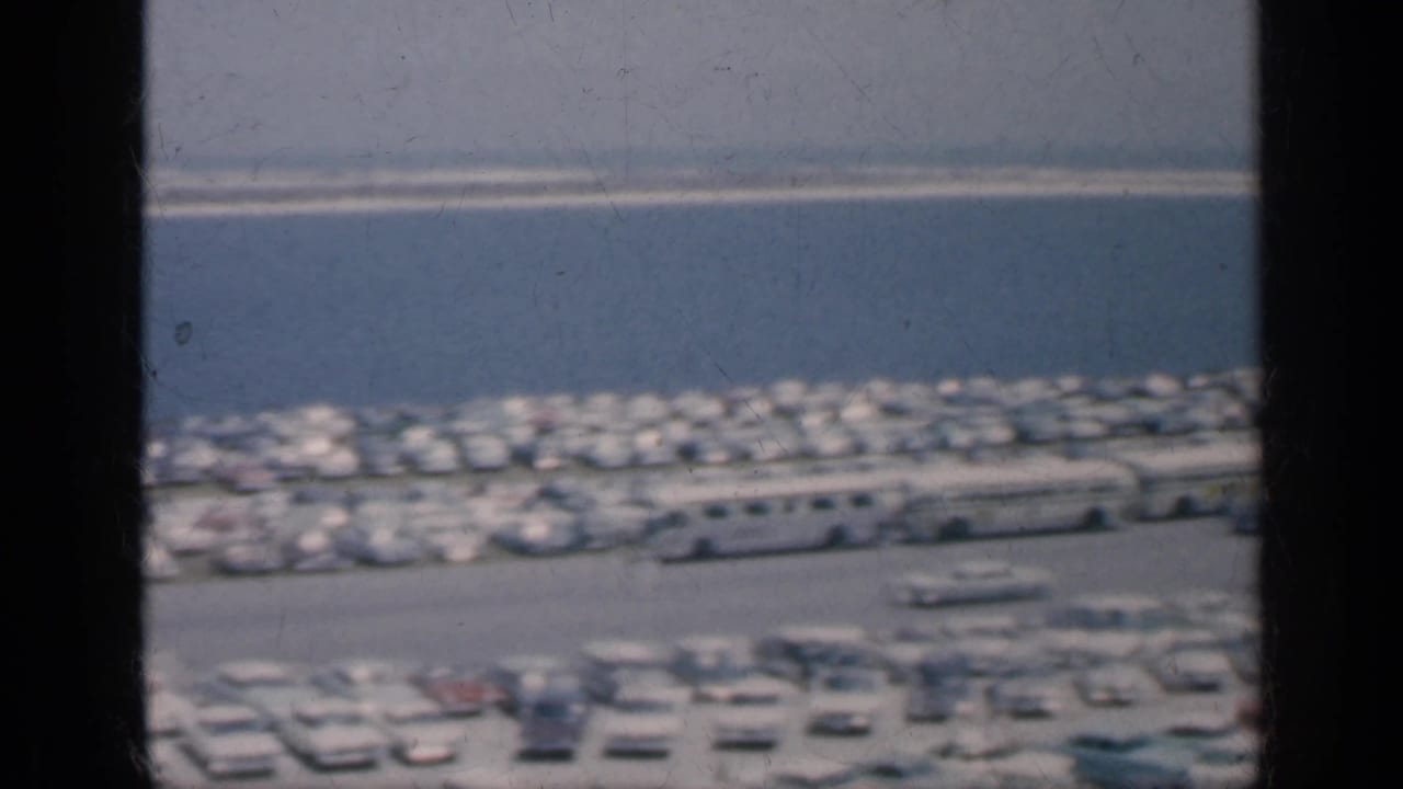 1962 Cuba home-movie frame showing a busy waterfront parking area, rows of cars, buses, and a low structure near the shoreline.