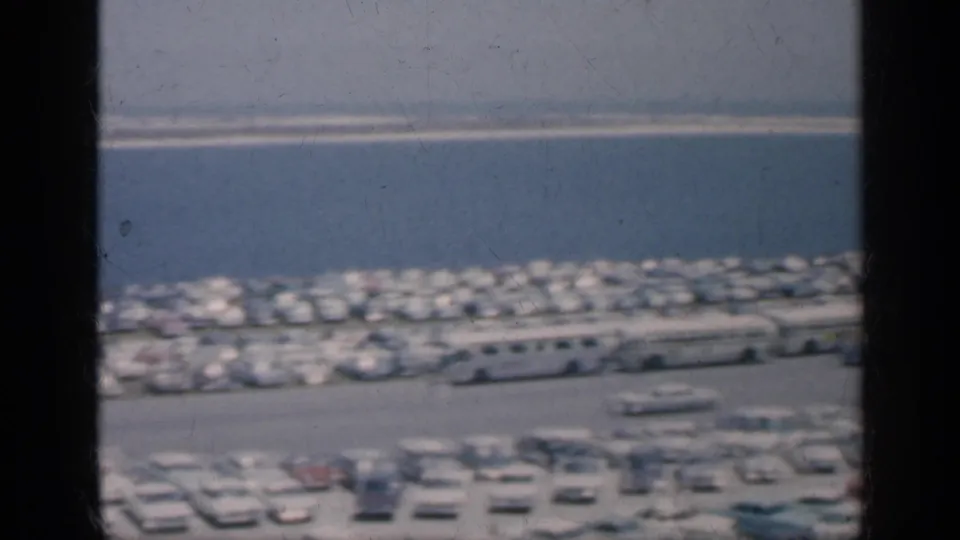 1962 Cuba home-movie frame showing a busy waterfront parking area, rows of cars, buses, and a low structure near the shoreline.