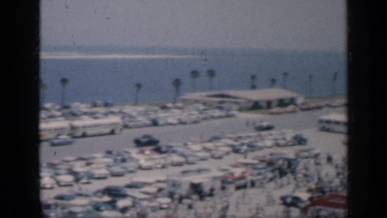 1962 Cuba travel footage frame with a waterfront parking lot, palm trees, a low building, rows of cars, and small distant figures.