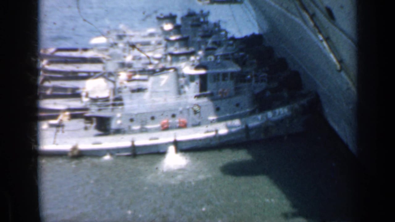 Harbor view of gray naval vessels beside a large aircraft carrier hull.