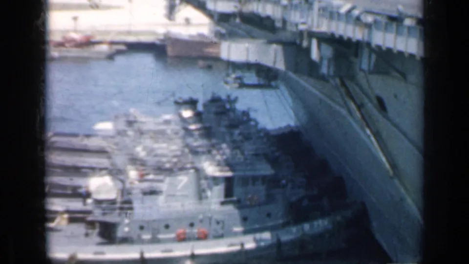 A moving 8mm view over a Cuban harbor shows an aircraft carrier flight deck, docked gray boats, pier structures, and surrounding water.