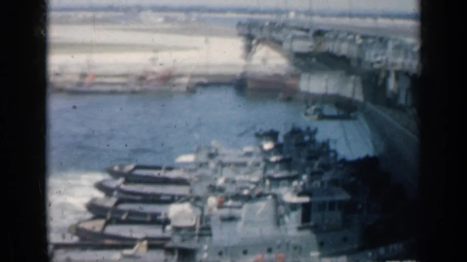 Soft 8mm frame from the Cuba harbor pan shows tugboats near a carrier deck, a pier in the background, and open harbor water.