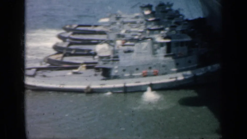 The final frame shows gray tugboats and naval craft alongside a large carrier hull, with harbor water and bright spray in the foreground.