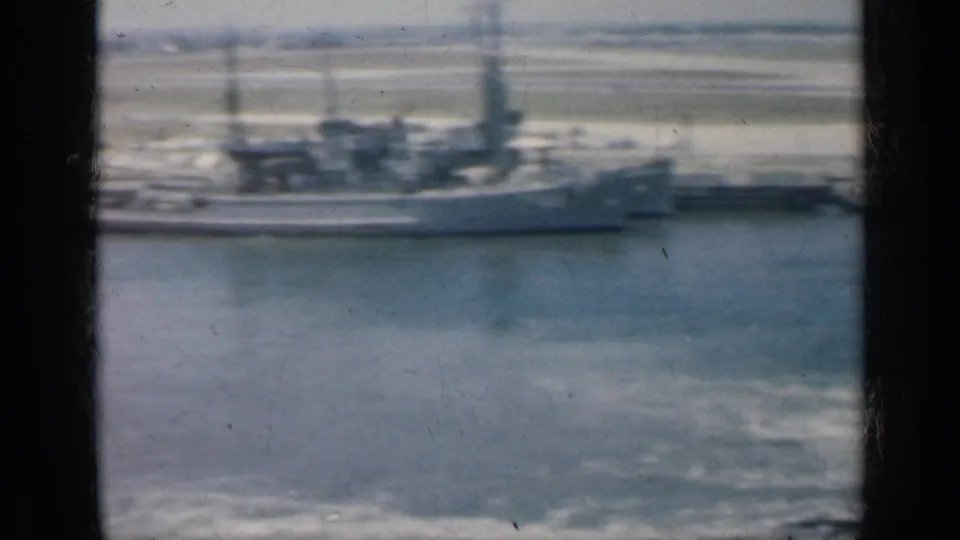 1962 Cuba home-movie frame with a long gray vessel along the waterfront, filmed from far across the harbor.