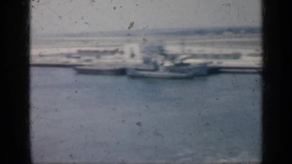 1962 Cuba 8mm frame showing a soft, distant view of boats and a larger gray ship beside the harbor shoreline.