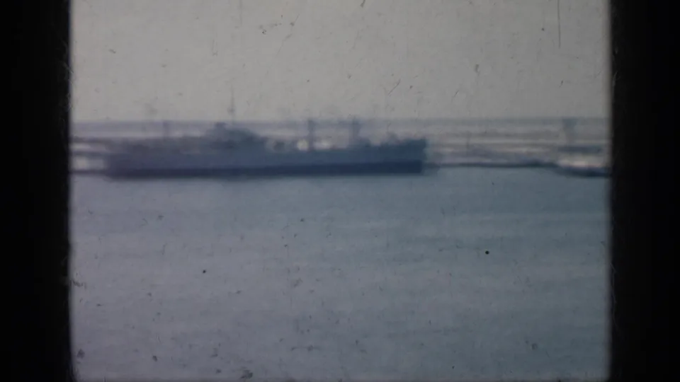 1962 Cuba 8mm frame showing a long gray vessel in profile across harbor water, with distant shoreline and heavy film wear visible.