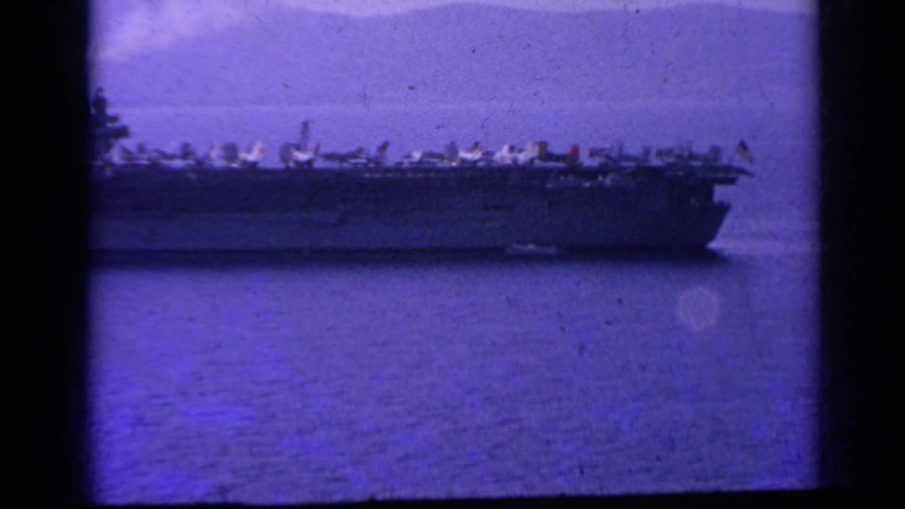 Distant aircraft carrier crossing calm water with jets lined along the deck.