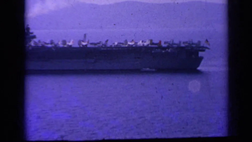 1962 Italy-associated 8mm frame of a distant U.S. Navy aircraft carrier on calm sea, with parked jets visible along the flight deck.