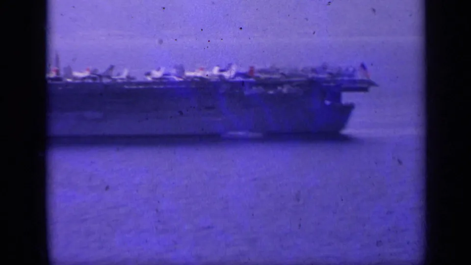 Side view of a large U.S. Navy carrier in the Italy-associated 1962 home-movie footage, with multiple jets lined on the flight deck.