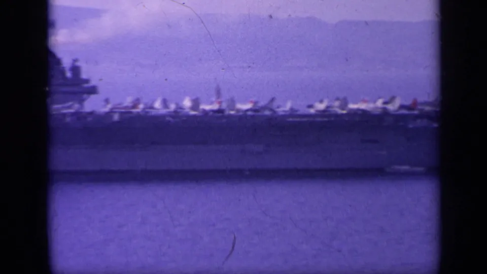Closer 8mm view of the carrier flight deck, showing numerous parked jets against calm water and a hazy land backdrop.