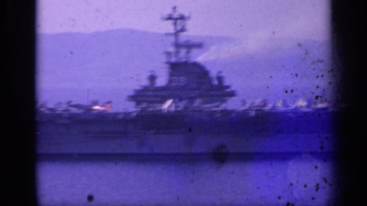 Distant aircraft carrier with island tower and jets on deck across calm water.
