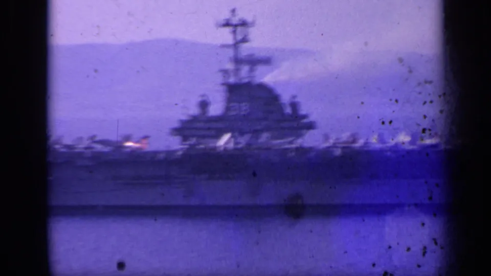 1962 Italy-associated 8mm frame showing a distant U.S. Navy aircraft carrier with deck aircraft, hazy hills, and purple film cast.