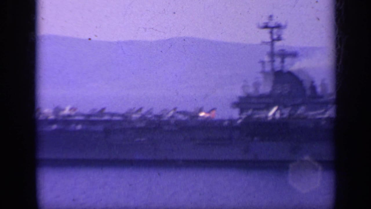 Soft 8mm view of a U.S. Navy carrier at sea, its deck aircraft silhouetted against hazy coastal hills.