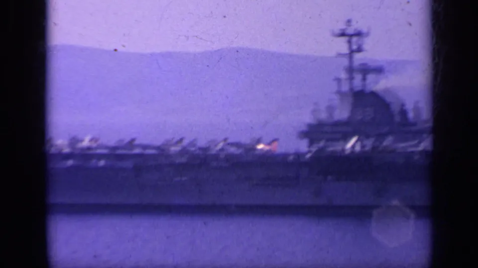 Soft 8mm view of a U.S. Navy carrier at sea, its deck aircraft silhouetted against hazy coastal hills.